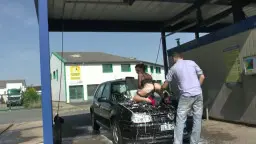 Geile Teen in schwarzem Netzbody beugt sich provokant über die Motorhaube eines schwarzen Autos, während ein Mann hinter ihr steht.