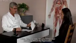 Grauhaariger Arzt im weißen Kittel sitzt am Schreibtisch und spricht mit brünetter Patientin im Praxiszimmer mit Anatomieposter.