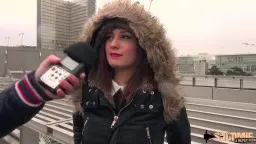 Junge Brünette mit Pelz-Kapuze und rotem Lippenstift blickt verführerisch in die Kamera vor modernem Hochhaus.