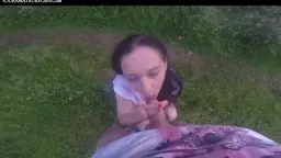 Junge Frau mit langen dunklen Haaren kniet auf bunter Decke im Gras und bläst einen dicken Schwanz mit vollem Einsatz.