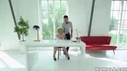 BBW Blondine in weißem Kleid beugt sich über einen Tisch, während ein junger Mann von hinten ihre Hüften packt und sie küsst.