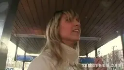 Nahaufnahme einer blonden Milf mit Ponyfrisur und weißen Rollkragenpullover auf einem Bahnsteig, die direkt in die Kamera spricht.