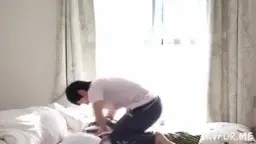 Japanerin in engem geringeltem Minirock beugt sich auf dem Bett lasziv nach vorne, während sie von einem Mann von hinten betrachtet wird.